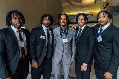Five Black male students in suits and ties pose together in the atrium of Goodwin Hall.